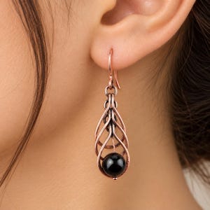 May include: A copper-toned metal dangle earring featuring a cage-like teardrop frame constructed from curved wire loops. A single, smooth, spherical black bead is suspended within the center of the wire structure, hanging from a simple hook.