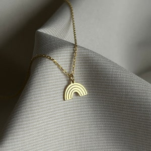 May include: A gold chain necklace with a small rainbow pendant. The necklace is on a white fabric background.