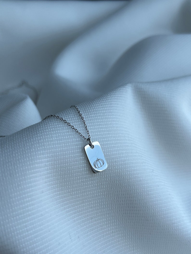May include: A silver necklace with a small rectangular pendant featuring a simple line drawing of a pumpkin.