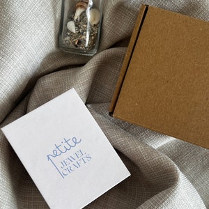 May include: A white box with the text "petite JEWEL CRAFTS" printed in blue. The box is sitting on a beige fabric surface. A small glass bottle filled with sand and seashells is also visible in the image.
