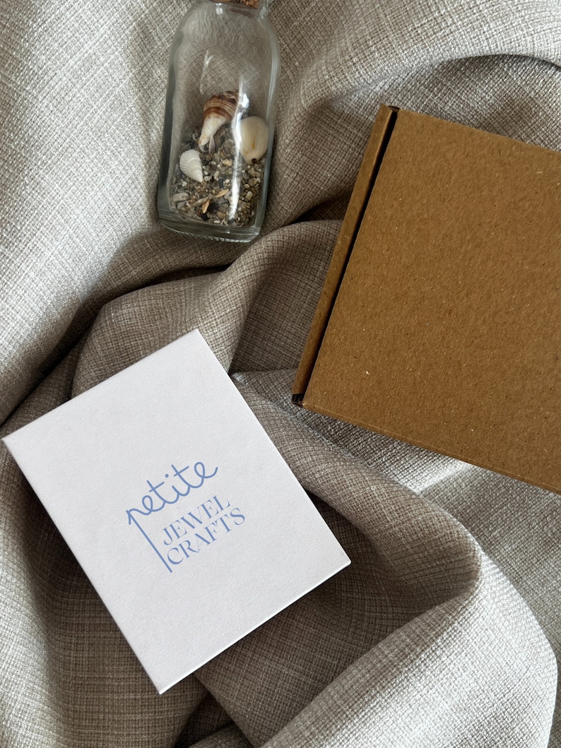 May include: A white box with the text "petite JEWEL CRAFTS" printed in blue. The box is sitting on a beige fabric surface. A small glass bottle filled with sand and seashells is also visible in the image.