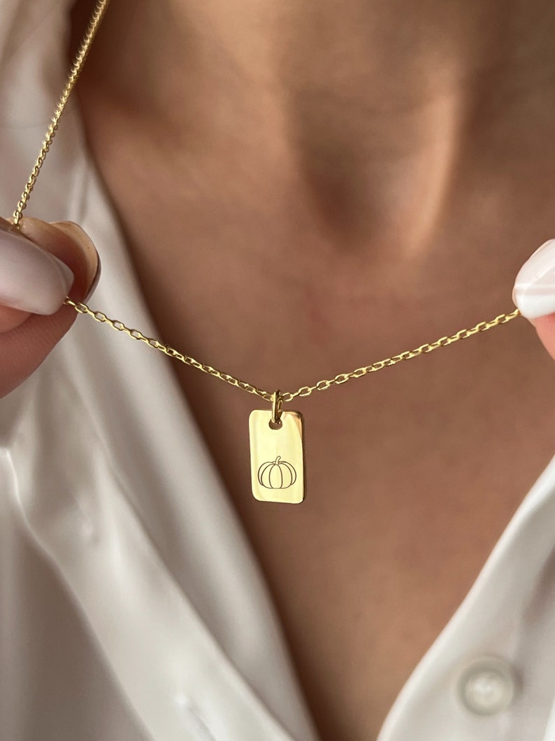 May include: A gold chain necklace with a rectangular pendant featuring a simple line drawing of a pumpkin.