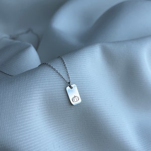 May include: A silver chain necklace with a small rectangular pendant featuring a simple line drawing of a pumpkin.