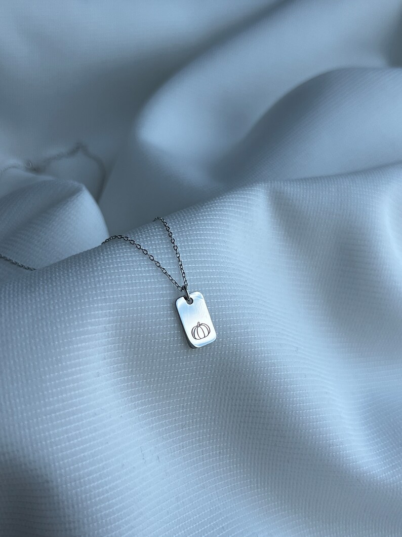 May include: A silver chain necklace with a small rectangular pendant featuring a simple line drawing of a pumpkin.