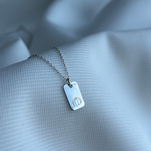May include: A silver necklace with a small rectangular tag pendant featuring a simple line drawing of a pumpkin.