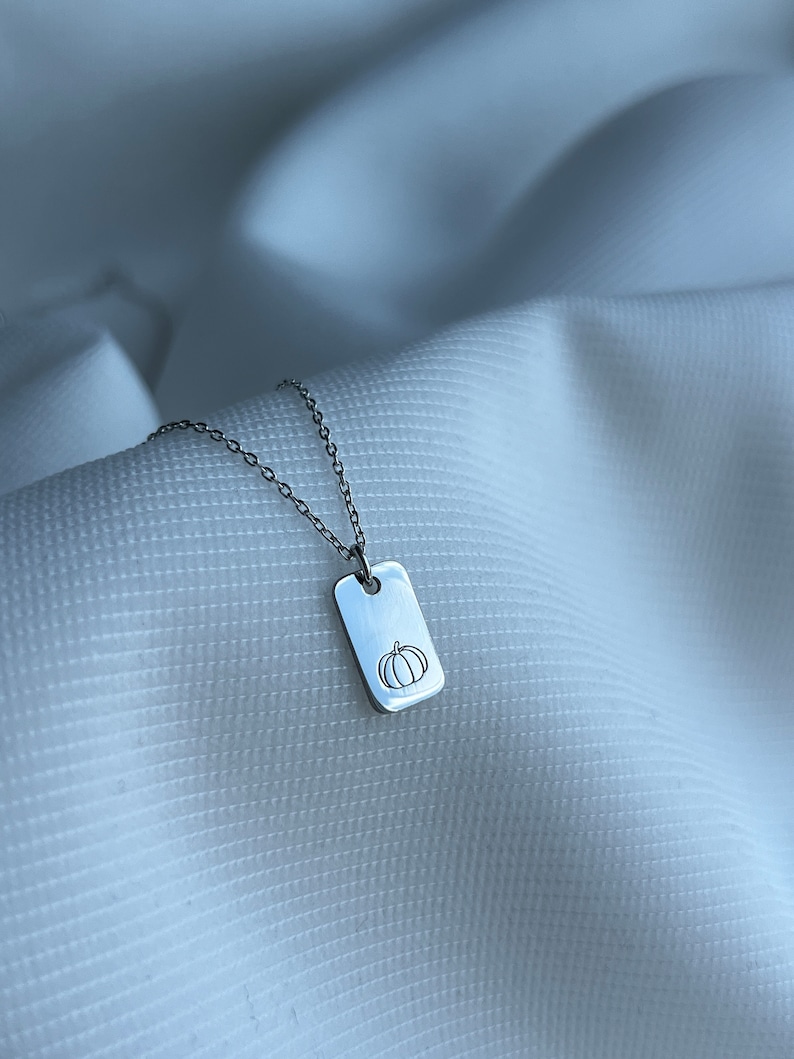 May include: A silver necklace with a small rectangular tag pendant featuring a simple line drawing of a pumpkin.