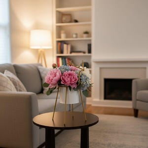 Puede incluir: Un arreglo floral en una maceta blanca con patas doradas, con peonías rosas e hortensias azules, se encuentra sobre una mesa redonda marrón oscuro. La mesa está en un salón con un sofá gris, una chimenea y una estantería.