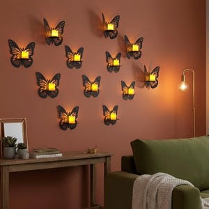 Op de afbeelding: Een verzameling zwarte metalen vlinder wandkandelaars, elk met een brandende kaars. De vlinders zijn geplaatst op een warm getinte muur, wat een decoratieve sfeer creëert. Een houten tafel en een groene bank staan op de voorgrond.