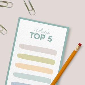 May include: A white card with a teal border and the text "today's TOP 5" written in green. There are five blank lines with checkboxes next to each line. A yellow pencil is lying on top of the card. Three silver paper clips are scattered around the card.
