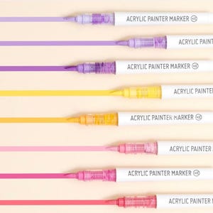 May include: Seven acrylic paint markers with white barrels and black lettering. The markers are arranged vertically with the tips facing down. Each marker has a different colour of paint visible in the tip, including purple, yellow, orange, pink, and red.