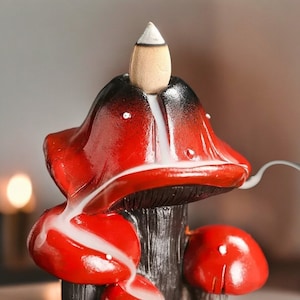 May include: Red ceramic incense burner shaped like a mushroom with a cone-shaped incense holder on top. The incense burner has a black base and white smoke is rising from the top.