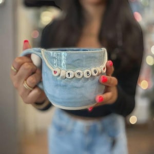 Könnte beinhalten: Hellblaue Keramiktasse mit weißem Henkel und Perlenbesatz. Die Perlenbuchstaben ergeben das Jahr "1989" mit herzförmigen Akzenten. Die Tasse hat eine strukturierte Oberfläche und einen hellblauen Innenraum.