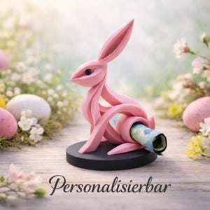 Puede incluir: Una escultura de conejo rosa con un ojo azul y un billete enrollado en su interior. El conejo está sobre una base circular negra. La palabra "Personalisierbar" está escrita en cursiva en la parte inferior.