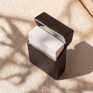 May include: A black leather box with a hinged lid, open to reveal a white rectangular object inside. The box is sitting on a textured, off-white surface, with shadows cast across it.