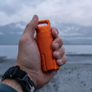 May include: An orange, cylindrical container with a textured surface and a loop at the top is held in a hand. The container is made of metal and appears to be waterproof. The background shows a body of water and mountains under a cloudy sky.