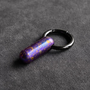May include: A purple capsule-shaped keychain with a celestial design of moons and stars in gold. The keychain is attached to a black metal ring. The capsule has a metallic sheen.