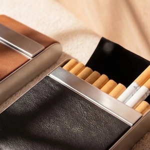 May include: Two cigarette cases, one brown and one black, with silver-colored metal accents. The black case is open, revealing several cigarettes. The cases have a leather-like texture and a rectangular shape.