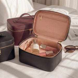 May include: A black makeup case with a gold zip, open to reveal a pink interior and beauty products. Two other cases, one black and one burgundy, are also visible. Tortoiseshell sunglasses rest nearby on a white surface.