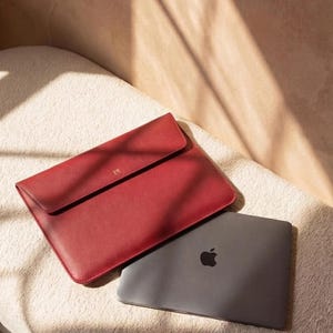 Peut inclure: Une housse d'ordinateur portable en cuir rouge avec une fermeture à rabat et des initiales dorées, à côté d'un ordinateur portable gris avec le logo Apple. La housse et l'ordinateur portable sont sur une surface texturée blanc cassé.