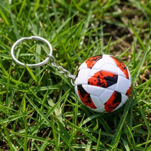 May include: A spherical soccer ball keychain featuring a white base with a repeating pattern of orange and black pixelated graphic shapes. The ball is attached to a silver-toned metal split ring by a short, flexible silver-toned metal chain link.
