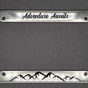 May include: A silver license plate frame with the text "Adventure Awaits" and a mountain range design at the bottom.