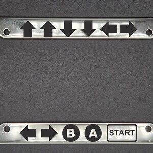 May include: A silver license plate frame with black arrow and button graphics. The graphics include four up arrows, two right arrows, one left arrow, and the words "B", "A", and "START".
