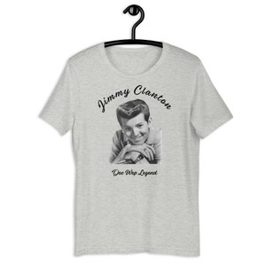 May include: A light gray t-shirt featuring a black and white image of Jimmy Clanton. The text "Jimmy Clanton" is arched above the image, and "Doo Wop Legend" is below. The shirt is hanging on a black hanger.