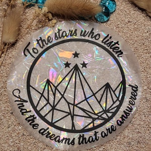Könnte beinhalten: Ein weißer, schimmernder Aufkleber mit einem schwarzen Kreis, der eine Bergkette mit drei Sternen darüber enthält. Der Text "To the stars who listen and the dreams that are answered" ist um den Kreis herum geschrieben.