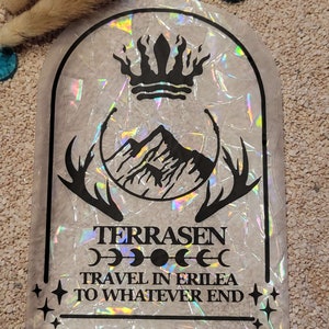 Puede incluir: Una pegatina holográfica con un diseño en blanco y negro. El diseño presenta una corona, montañas, cuernos de ciervo y el texto "TERRASEN TRAVEL IN ERILEA TO WHATEVER END".