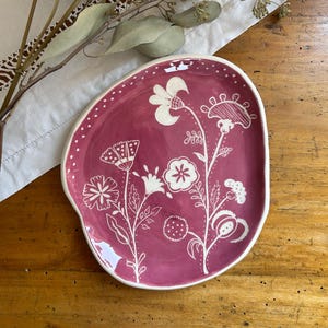 Puede incluir: Un plato de cerámica ovalado hecho a mano con una base magenta oscuro y un borde blanco irregular. El plato presenta un diseño floral en blanco, con varias flores y hojas estilizadas. El plato está sobre una superficie de madera.