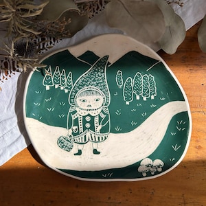 Può includere: Piatto in ceramica con una scena boschiva stravagante. Il piatto presenta uno sfondo verde con un sentiero bianco e una figura con un cappuccio. Sono raffigurati anche alberi e montagne. Il piatto ha un diametro di circa 25 cm.