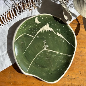 Può includere: Un piatto in ceramica color bianco sporco di forma rotonda con un bordo irregolare e organico. L'interno presenta un paesaggio dipinto a mano in verde scuro con un disegno a linee bianche di una piccola capanna, montagne, una luna crescente e stelle.