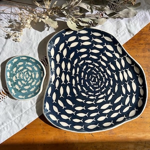 May include: Two ceramic plates with a fish design. The larger plate is navy blue with white fish arranged in a spiral pattern. The smaller plate is teal with a similar fish design. Both plates have a white border.