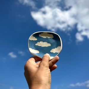 Peut inclure: Un petit plat en céramique fait à la main avec un intérieur bleu et des motifs de nuages blancs. Le plat est tenu contre un ciel bleu vif avec des nuages blancs et duveteux. Le plat est une œuvre d'art unique.