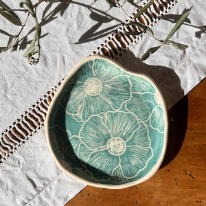 Puede incluir: Un plato de cerámica pequeño, de forma irregular, con interior turquesa y un estampado floral blanco. El borde es de color blanco crema. El plato está sobre un paño blanco con un ribete de encaje, con ramas de olivo al fondo.