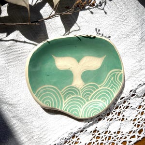 Pode incluir: Um pequeno prato de cerâmica de forma irregular, feito à mão, com um esmalte verde claro liso. O centro apresenta uma cauda de baleia esculpida em tom creme, sem esmalte, que emerge sobre padrões de ondas concêntricas gravados na argila.