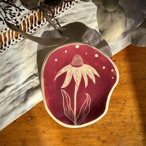 Könnte beinhalten: Eine handgefertigte Keramikschale mit tiefrotem Innenraum und weißem Blumendesign. Die Schale hat eine unregelmäßige Form und einen cremefarbenen Rand. Weiße Punkte sind um das Blumendesign verstreut. Die Schale steht auf einer Holzoberfläche.