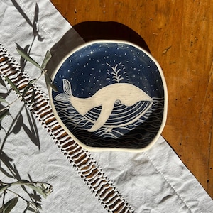 Può includere: Un piatto in ceramica rotondo e modellato a mano con un bordo irregolare e organico. L'interno presenta uno smalto blu navy profondo con un'illustrazione a sgraffio bianca di una balena che nuota in acqua ondulata, con un getto d'acqua.