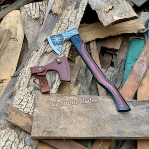 May include: A handcrafted axe with a black and red handle and a silver blade featuring a snowflake design. Includes a brown leather sheath. The axe is displayed on a wooden surface, with the text "Jak Smith Blades".