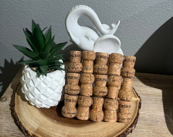 Op de afbeelding: Een decoratieve opstelling met champagnekurken gestapeld in een toren, een wit keramisch olifantenbeeldje en een kleine vetplant in een witte geometrische pot. De items zijn geplaatst op een houten schijf.