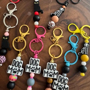 May include: Five colorful keychains with different charms, including a black cat, a skeleton, and a dog paw print. Each keychain has a silver, gold, or black metal ring and a variety of colorful beads.