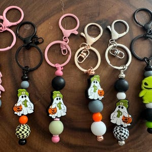 May include: Six Halloween-themed keychains with different characters and colors. Each keychain features a ghost, a green monster, and a pink monster with a black and white checkered bead, a green bead, and a white bead.