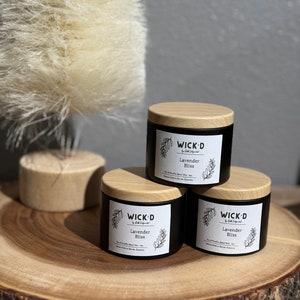 May include: Three black candles with wooden lids are displayed on a wooden slab. Each candle features a white label with the text "WICK'D Lavender Bliss" and a botanical illustration. A feathery plant is in the background.