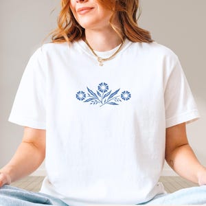 May include: White t-shirt with a blue floral design. The graphic features three stylized flowers with leaves and stems. The text "PREMIUM UNISEX TEE" is printed in a black, bold font at the bottom of the image.