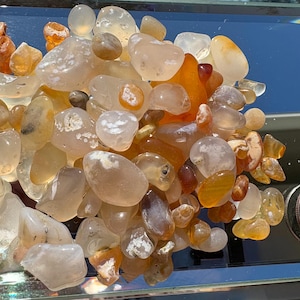 May include: A collection of polished, tumbled gemstones in shades of orange, yellow, and clear. The stones vary in size and shape, with a quarter coin for size comparison. The gemstones are displayed on a reflective surface.