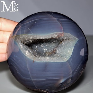 Puede incluir: Una esfera de ágata azul oscuro pulida que presenta una cavidad de geoda horizontal central llena de pequeños cristales translúcidos y brillantes. La superficie lisa de la piedra muestra sutiles patrones de bandas concéntricas en tonos azul marino y pizarra.