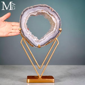 Könnte beinhalten: Eine kreisförmige Achat-Geodenscheibe mit hohler Mitte und kristallinem Inneren, die konzentrische Bänder in Grau, Weiß und durchscheinendem Violett aufweist. Die Geode ist auf einem goldfarbenen Metallständer mit V-förmigem Rahmen montiert.