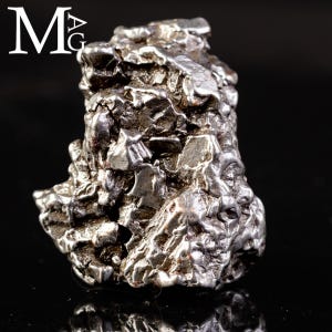 May include: A metallic, silver-toned iron meteorite specimen with a highly irregular, craggy surface texture featuring deep crevices and raised ridges. The text "MAG" is printed in white in the upper left corner against a dark background.