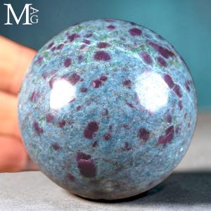 May include: A polished spherical stone featuring a textured surface with a mottled blue and green base colour, accented by numerous embedded deep magenta and dark red crystalline inclusions throughout the entire rounded mineral form.