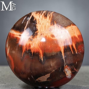 Puede incluir: Una piedra esférica pulida con una superficie lisa y brillante, con patrones de color marrón oscuro, negro y rojo anaranjado vibrante que se asemejan a llamas fluidas o vetas de madera orgánica. La esfera tiene un acabado de alto brillo.